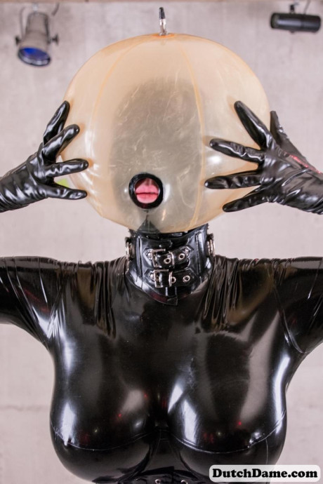 Latex attired slave is affixed with an inflatable hood in dungeon setting